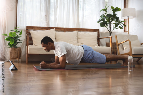 Side view of Black African American man looking at digital tablet and doing plank exercise at home. Wellness, Fitness and healthy lifestyle. photo