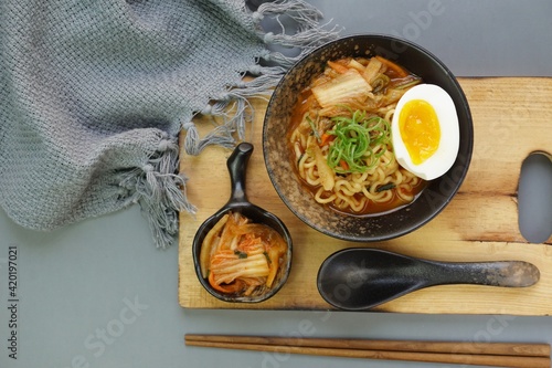 Top view of instan ramyeon or Korean noodles soup on black bowl, topped with half boiled egg, leek and Kimchi on small bowl. Isolated gray background with spoon, napkin and wooden board