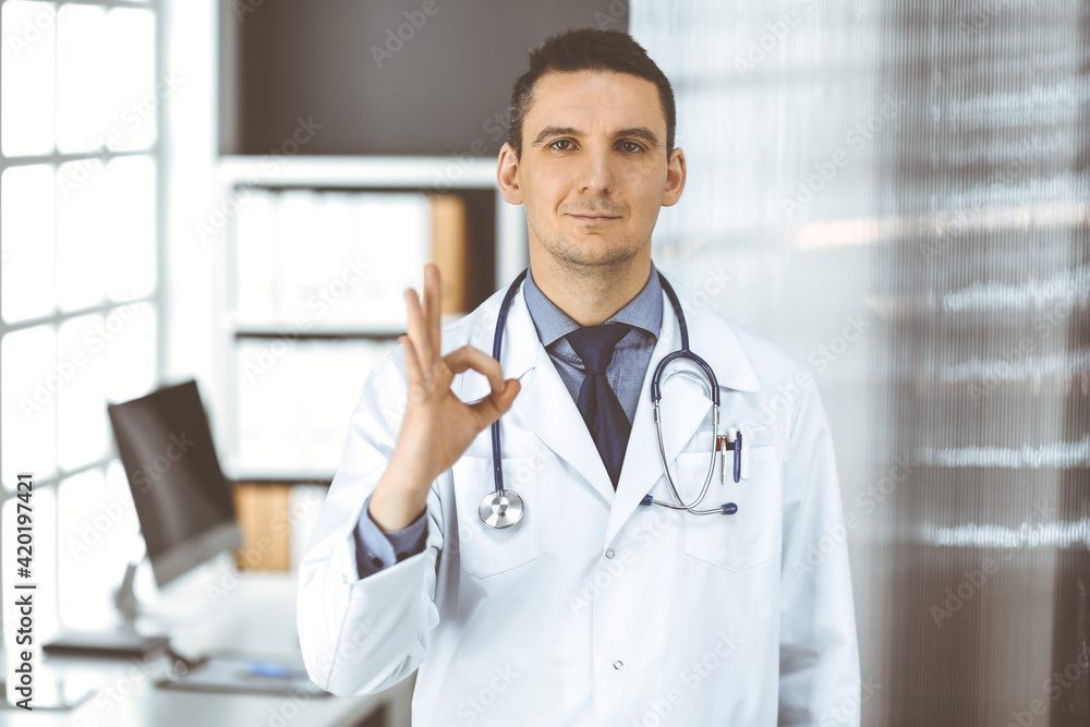 Friendly male doctor standing straight with OK sign in clinic near his working place, closeup. Perfect medical service in hospital. Medicine and healthcare concept