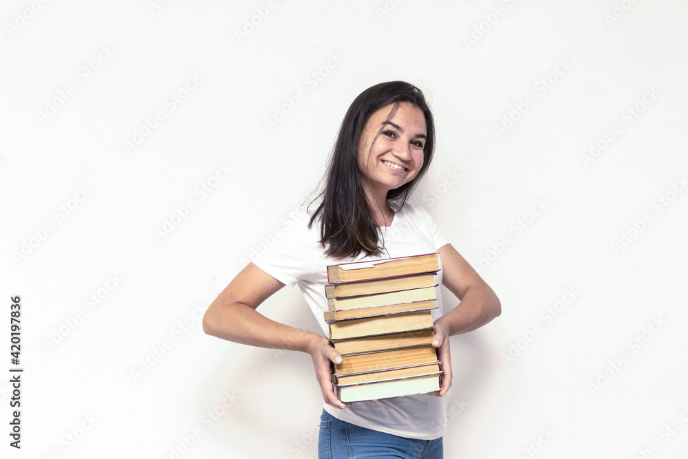 Young smiling student woman in white t-shirt holding textbooks and books in a pile on a white backdrop. College education concept. Positive emotions, facial expression Young smiling student woman in white t-shirt holding textbooks and books in a pile on a white backdrop. College education concept. Positive emotions, facial expression