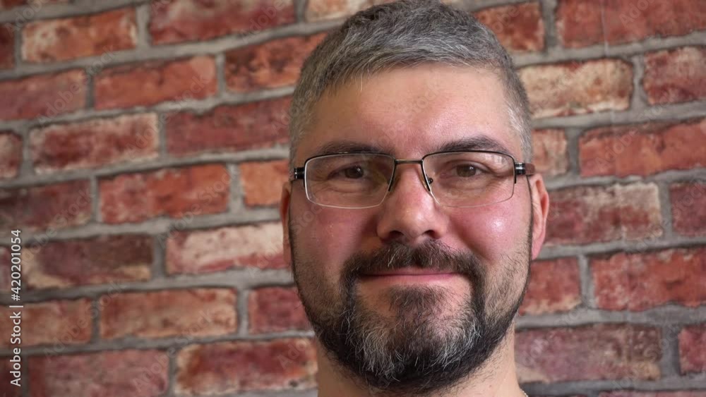 Bearded man in stylish glasses. Looks straight. Short haircut. Blue T-shirt. Poor eyesight. Against the background of a brick wall. Close-up. Bearded man in stylish glasses. Looks straight. Short haircut. Blue T-shirt. Poor eyesight. Against the background of a brick wall. Close-up.