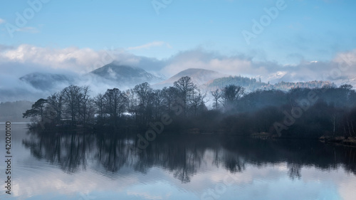 Dramatic landscape image looking across Derwentwater in Lake District towards Catbells snowcapped mountain with thick fog rolling through valley