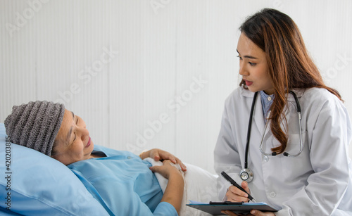 Young and beautiful doctor asking and note for diagnosis with Older Asian woman patient covered the head with clothes effect from chemo treatment in cancer cure process