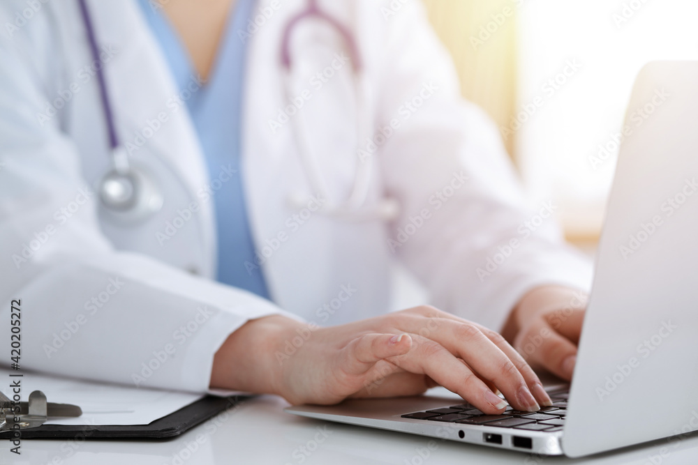 Unknown woman-doctor typing on laptop computer while sitting at the table in sunny clinic Unknown woman-doctor typing on laptop computer while sitting at the table in sunny clinic
