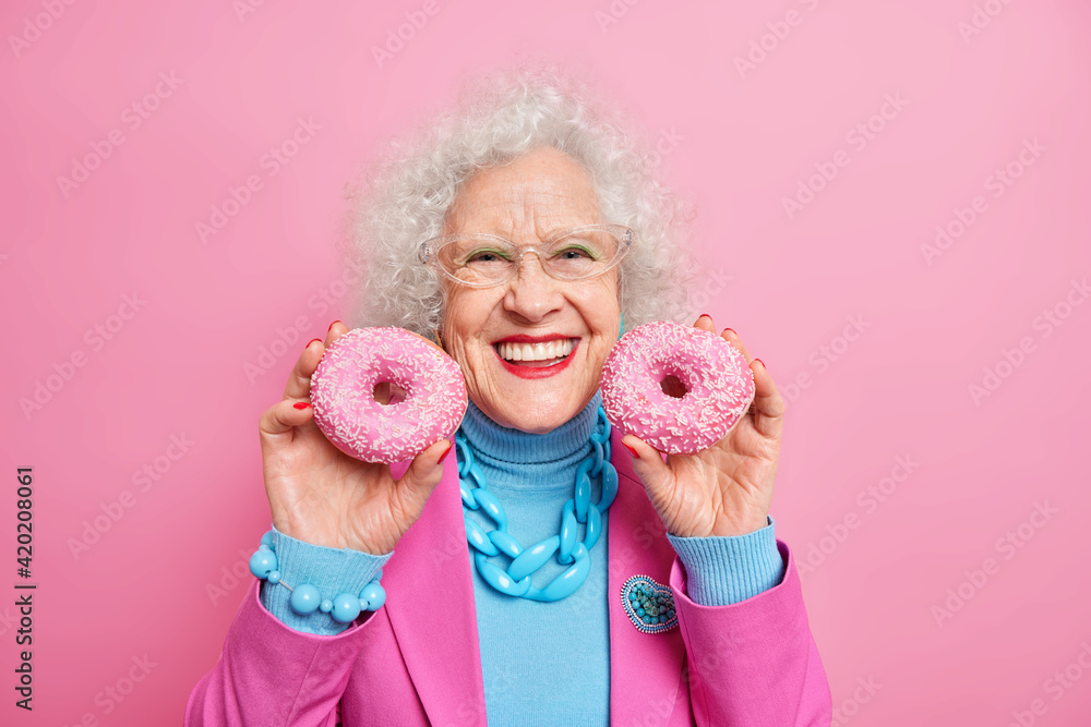 Positive beautiful wrinkled elederly European woman holds two delicious glazed doughnuts smiles broadly has good mood applies makeup fashionable clothes and jewelry isolated over pink background.