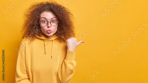 Attractive surprised curly haired woman keeps lips rounded points thumb away makes her choice on shopping dressed in casual hoodie poses over yellow background shows logo or promo deal on copy space © wayhome.studio 
