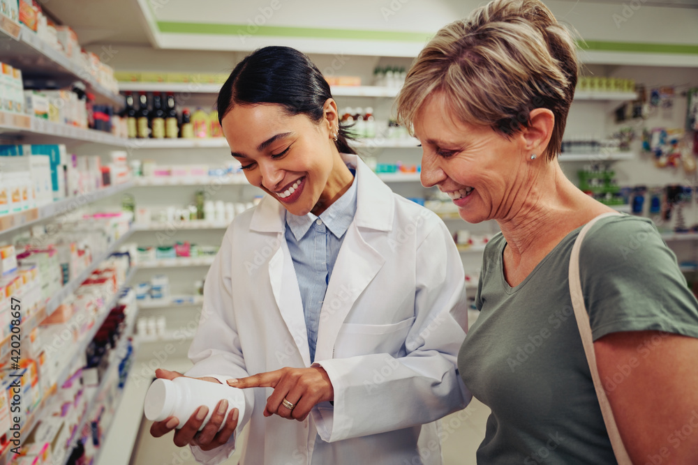 Smiling senior customer with pharmacist looking for medicinal details on bottle standing near shelves Smiling senior customer with pharmacist looking for medicinal details on bottle standing near shelves