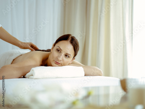 Pretty brunette woman enjoying treatment with hot stones in spa salon. Beauty concept