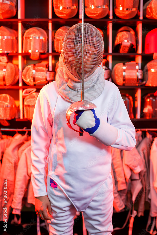 Young guy in a fencing suit with a sword in his hand, on a red background, neon light. The athlete trains. Sports, youth, healthy lifestyle.