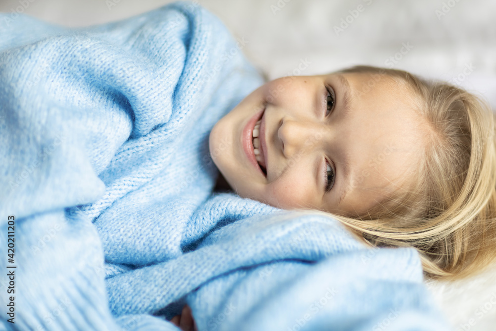 Cute little caucasian girl seven years old with blonde hair lying on bed at home in bedroom. Kid wearing stylish cozy knitted sweater blue color. Happy Child smiling Cute little caucasian girl seven years old with blonde hair lying on bed at home in bedroom. Kid wearing stylish cozy knitted sweater blue color. Happy Child smiling
