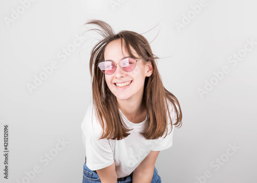 Brunette woman with hair fluttering in the wind, in pink glasses, a white t-shirt and jeans, smiling, looking at the camera on a gray background