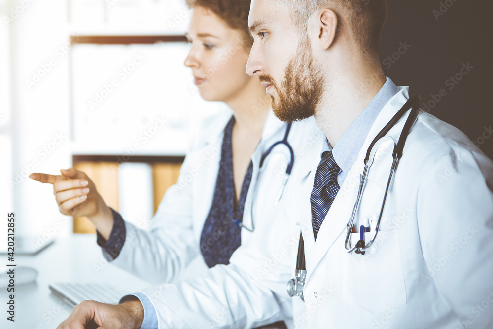 Red-bearded doctor with female colleague discussing current disease therapy while sitting at working place in sunny clinic. Team work in medicine Red-bearded doctor with female colleague discussing current disease therapy while sitting at working place in sunny clinic. Team work in medicine