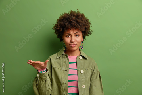 Dissatisfied dark skinned Afro American woman dressed in fashionable clothes shows something not very tall being not satisfied with size isolated over vivid green background. Hmm its so short