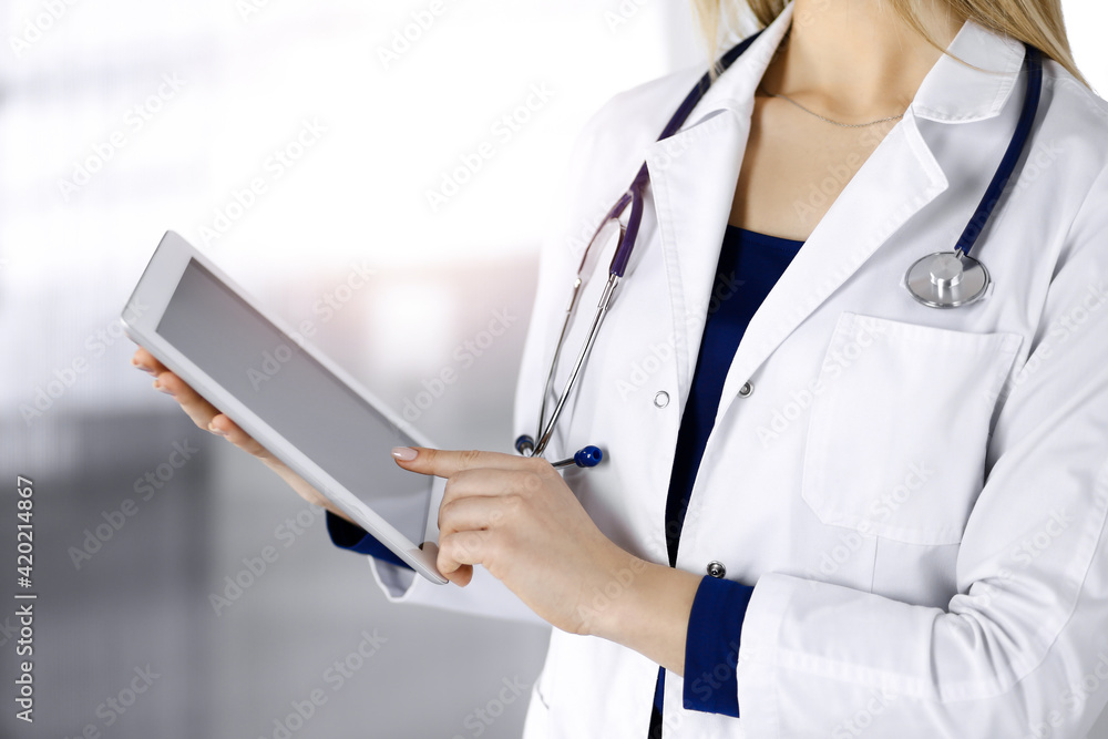 Unknown woman-doctor is holding a tablet computer in her hands, while standing in a sunny clinic cabinet. Female physician at work, close-up. Perfect medical service in a hospital. Medicine concept Unknown woman-doctor is holding a tablet computer in her hands, while standing in a sunny clinic cabinet. Female physician at work, close-up. Perfect medical service in a hospital. Medicine concept