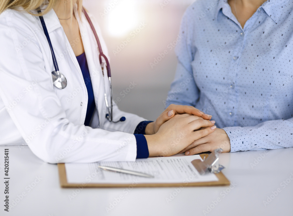 Unknown woman-doctor is holding her patient's hands to reassure a patient, while sitting together at the desk in the sunny cabinet in a clinic. Female physician is using a clipboard and a stethoscope Unknown woman-doctor is holding her patient's hands to reassure a patient, while sitting together at the desk in the sunny cabinet in a clinic. Female physician is using a clipboard and a stethoscope