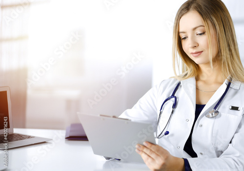 Young optimistic woman-doctor is using a clipboard, while sitting at the desk in her sunny cabinet in a clinic. Portrait of friendly female physician woman with a stethoscope. Perfect medical service