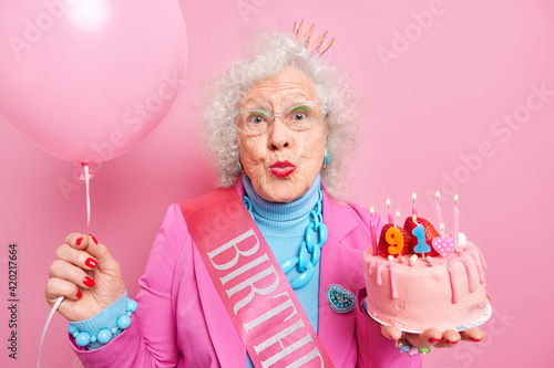 Lovely curly haired grandmother keeps lips folded wants to kiss someone acceps congratulations on her birthday poses with tasty cake and inflated balloon wears spectacles festive outfit poses indoor