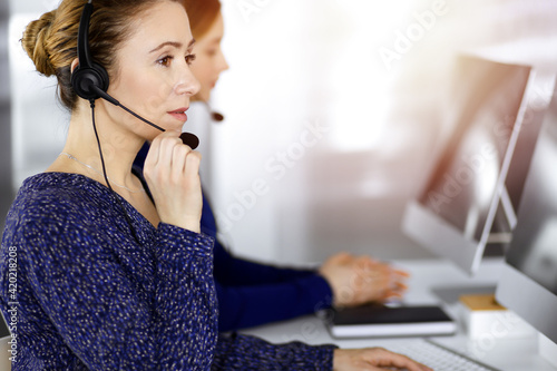 Two busineswomen have conversations with the clients by headsets, while sitting at the desk in a sunny modern office. Diverse people group in a call center. Telemarketing and customer service