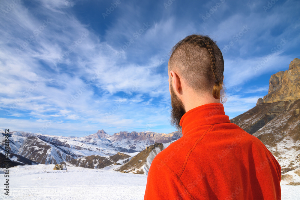 A bearded man with a stylish haircut in a red sweater stands with his back and looks into the mountains and rocks covered with snow