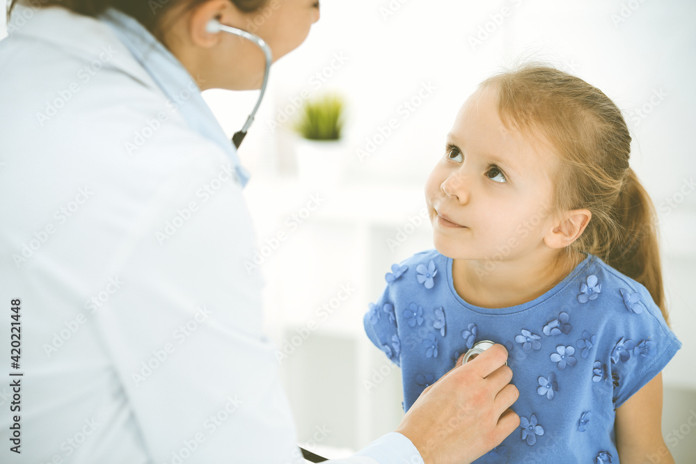 Doctor examining a child by stethoscope. Happy smiling girl patient dressed in blue dress is at usual medical inspection. Medicine concept Doctor examining a child by stethoscope. Happy smiling girl patient dressed in blue dress is at usual medical inspection. Medicine concept