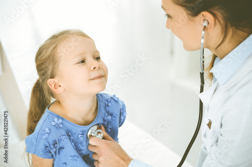 Doctor examining a child by stethoscope. Happy smiling girl patient dressed in blue dress is at usual medical inspection. Medicine concept