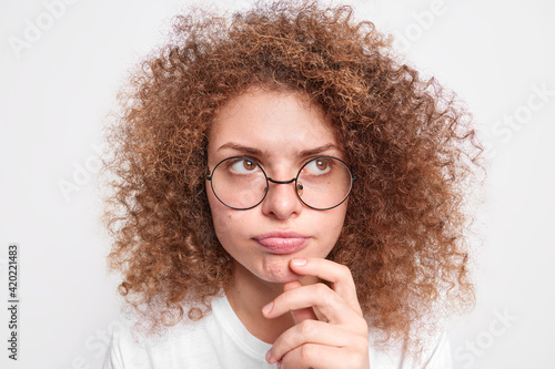 Close up shot of pensive thoughtful young woman with curly hair imagines something or plans makes assumptions wears round spectacles for vision correction poses indoor has some doubts stands sad