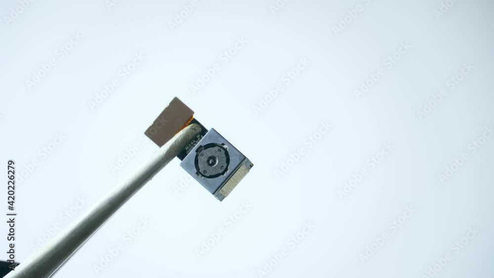 Close-up on the mobile phone camera module. Close-up tweezers or clip holds smartphone lens in studio shot white white background Close-up on the mobile phone camera module. Close-up tweezers or clip holds smartphone lens in studio shot white white background