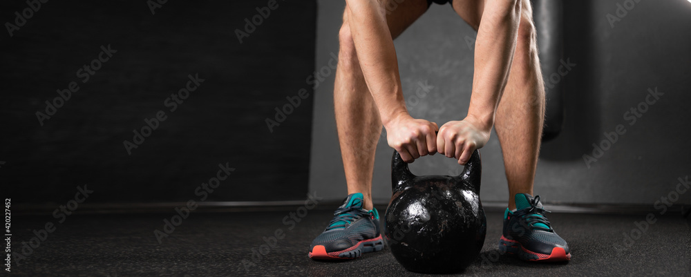 Male athlete in the gym lifts a heavy kettlebell with both hands. Legs and arms of a sports man on the right and place for text. - obrazy, fototapety, plakaty 