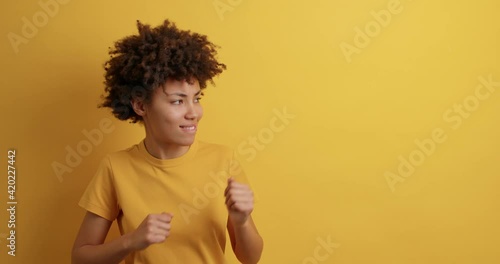 Positive curly haired woman shakes fists dances carefree dressed in casual clothes feels energetic moves with rhythm of music dressed casually poses against yellow background blank space for promo
