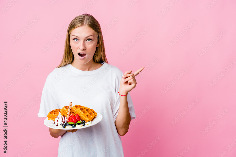 Young russian woman eating a waffle isolated pointing to the side