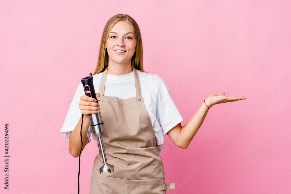 Young russian cook woman holding an electric mixer isolated makes scale with arms, feels happy and confident. Young russian cook woman holding an electric mixer isolated makes scale with arms, feels happy and confident.