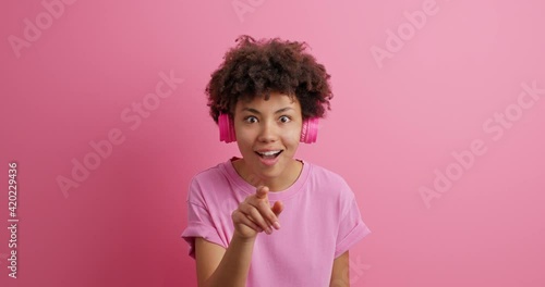 Wow how icredible it is. Impressed curly haired young woman indicates in front says wow wears casual t shirt wireless headphones on ears notices something amazing isolated over pink background photo