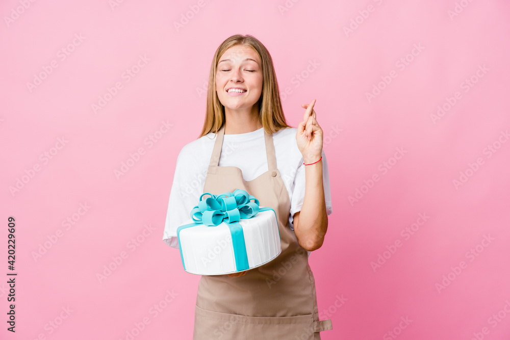 Young russian baker woman holding a delicious cake crossing fingers for having luck Young russian baker woman holding a delicious cake crossing fingers for having luck