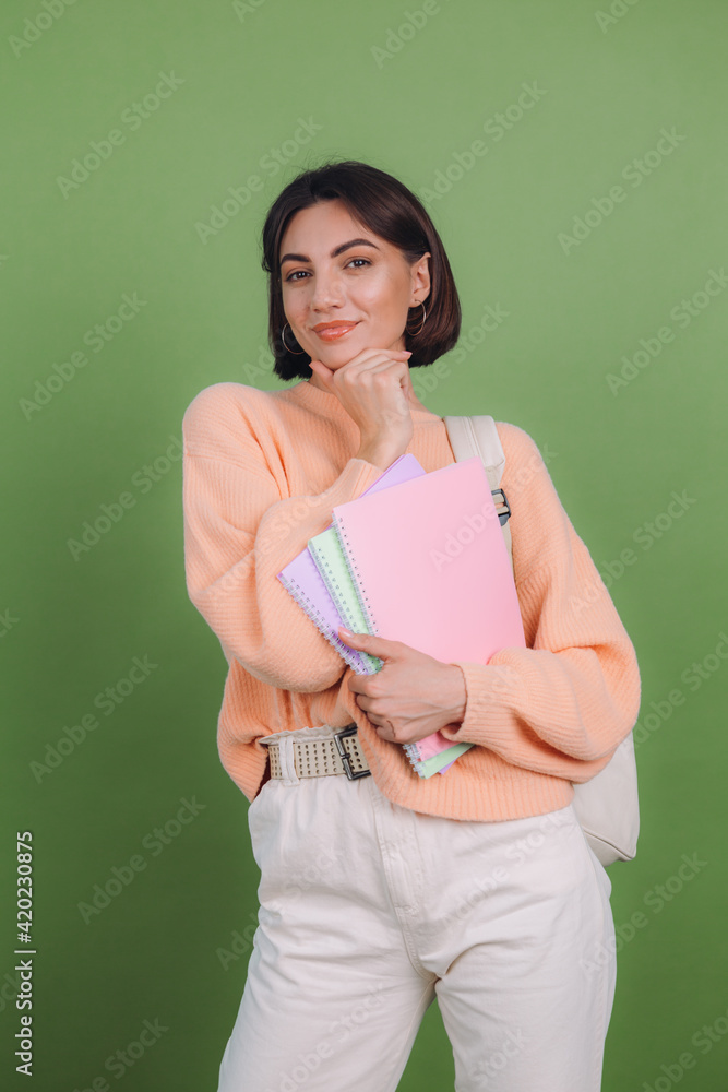 Young woman in casual peach sweater isolated on green olive color background with notebooks thoughtful positive touch chin with hand, education high school concept Young woman in casual peach sweater isolated on green olive color background with notebooks thoughtful positive touch chin with hand, education high school concept