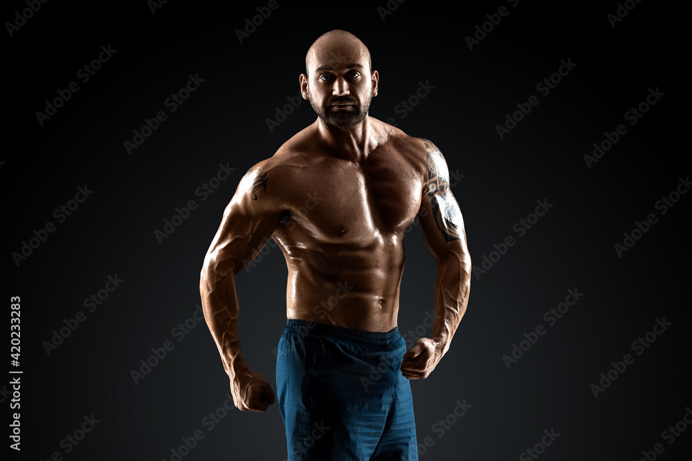 Naklejka premium Male bodybuilder with light stubble and bare torso shows muscularity against a dark background. The concept of a fitness club, doing sports, weightlifting. Copy space.