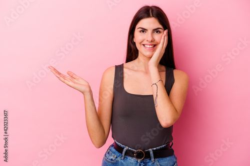 Young caucasian woman isolated on pink background holds copy space on a palm  keep hand over cheek. Amazed and delighted.