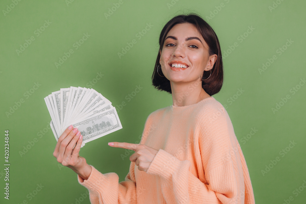 Young woman in casual peach sweater isolated on green olive background lucky holding fan of 100 dollar bills being excited smile and pointing with index finger Young woman in casual peach sweater isolated on green olive background lucky holding fan of 100 dollar bills being excited smile and pointing with index finger