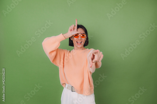 Young woman in casual peach sweater and orange glasses isolated on green olive background pointing at you mockery fool day showing horn symbol copy space