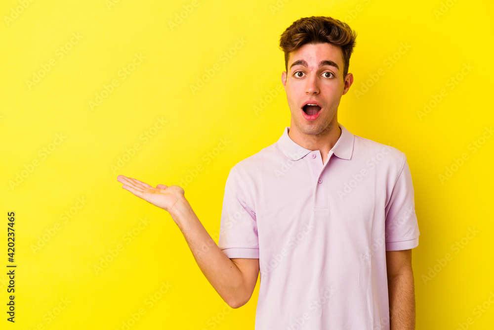 Young caucasian man isolated on yellow background impressed holding copy space on palm. Young caucasian man isolated on yellow background impressed holding copy space on palm.