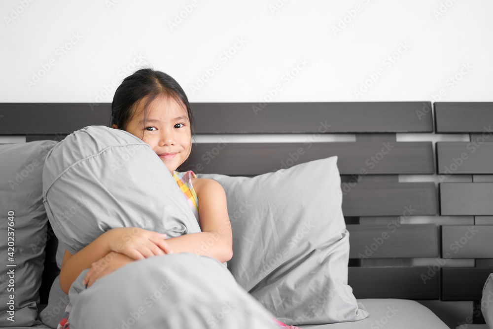 Asian child or kid girl sitting hugging soft bolster and leaning gray pillow on bed head and happy smile to comfortable relax on wake up bright after sleep in morning and bedroom at home for stay safe Asian child or kid girl sitting hugging soft bolster and leaning gray pillow on bed head and happy smile to comfortable relax on wake up bright after sleep in morning and bedroom at home for stay safe