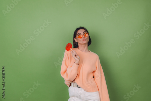 Young stylish woman in casual peach sweater and orange glasses isolated on green olive background with orange lollipop thoughtful look aside thinking copy space