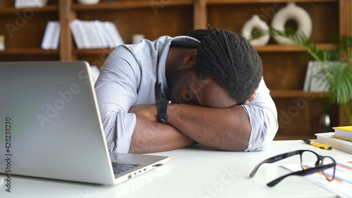 Exhausted African-American male office employee in smart casual wear is taking a nap on the workplace, feels tired and burnout, a multiracial man sleeps on the desk. Owerworked concept photo