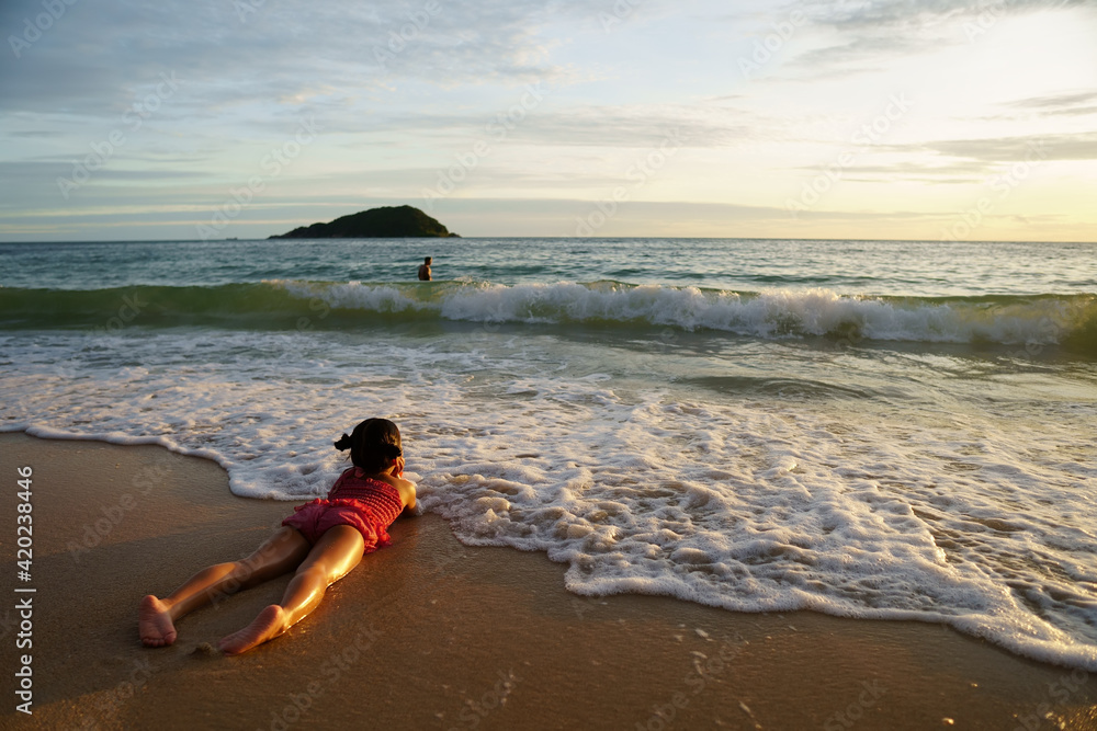 Obraz premium asian child happy or kid girl lie down on stomach hand to chin on island sand beach and sea with family on golden warm sunlight to morning sunrise or sunset for vacation travel on summer holiday relax