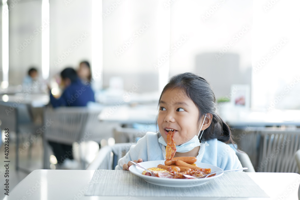 Asian child smile or kid girl wearing face mask under chin to enjoy eating delicious food by grilled bacon sausage on white dish with happy lunch or breakfast in morning at restaurant or food court Asian child smile or kid girl wearing face mask under chin to enjoy eating delicious food by grilled bacon sausage on white dish with happy lunch or breakfast in morning at restaurant or food court