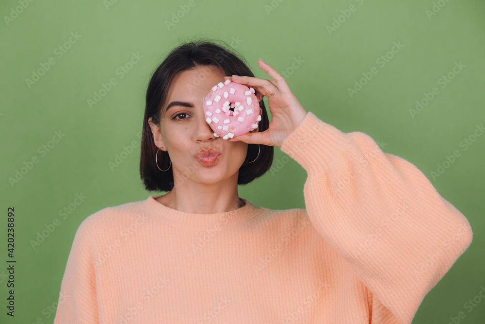 Young stylish woman in casual peach sweater  isolated on green olive background send kiss with pink donut copy space