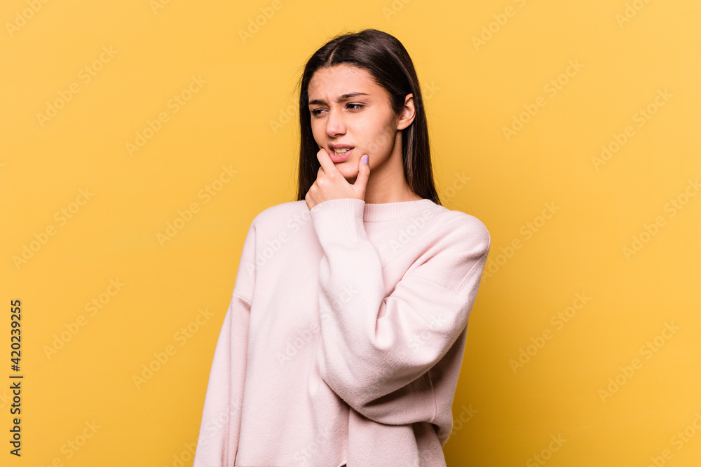 Young Indian woman isolated on yellow background touching back of head, thinking and making a choice.