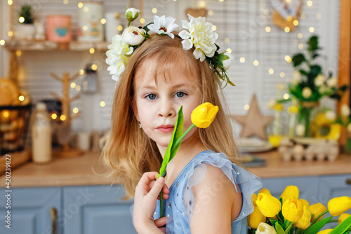 A little girl with a flower wreath on her head and a yellow tulip in her hand in the kitchen.
