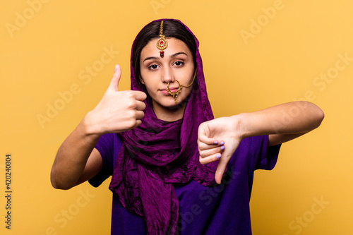 Young Indian woman wearing a traditional sari clothes isolated on yellow background showing thumbs up and thumbs down, difficult choose concept © Asier