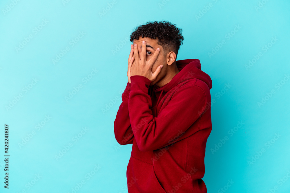 Young african american curly man isolated on blue blink through fingers frightened and nervous. Young african american curly man isolated on blue blink through fingers frightened and nervous.