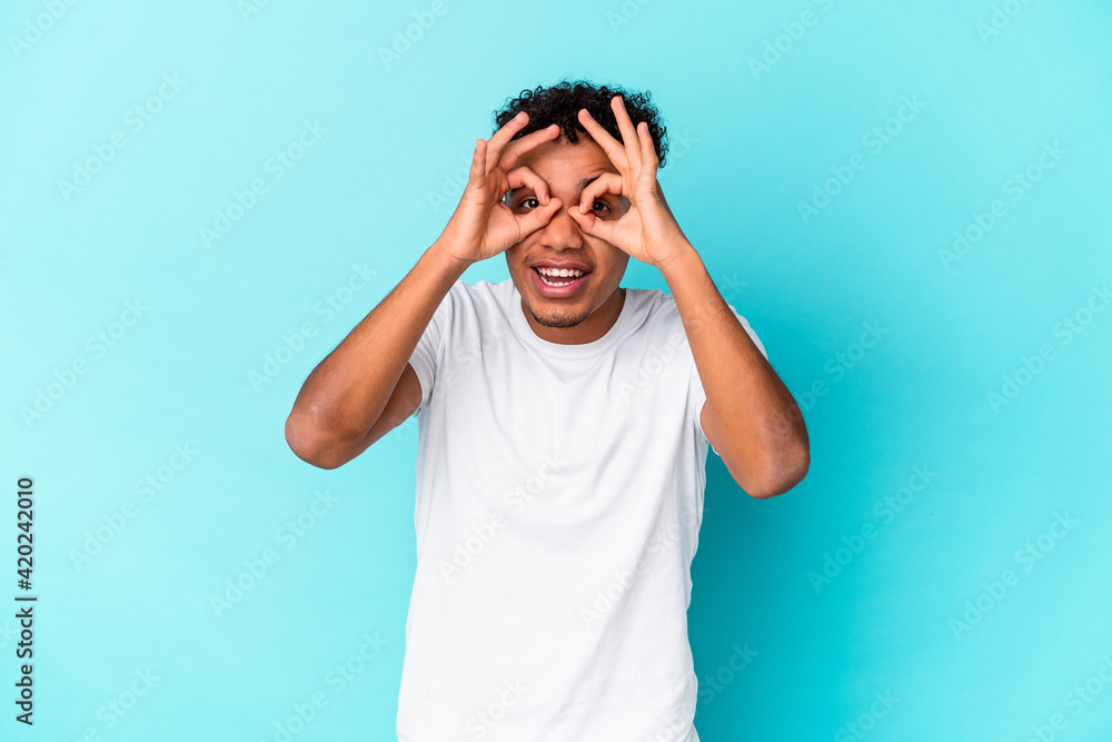 Young african american curly man isolated on blue showing okay sign over eyes Young african american curly man isolated on blue showing okay sign over eyes